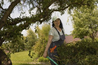 Çizgili tişört ve gri tulumlu gülümseyen kadın bahçıvan yeşil bir arka bahçede budanmış dallar topluyor. Mevsimlik bahçe bakımı, budanma ve sürdürülebilir yaşam kavramı.