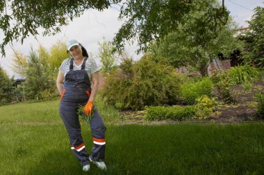 Çizgili gömlekli ve iş tulumlu bir kadın bahçede dikiliyor ve bir elinde kesik dallar tutuyor. Bahçe işleri, açık hava işleri ve mevsimlik bahçe temizliği kavramı.