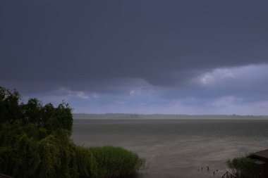 Büyük bir göl üzerinde sağanak yağış, üzerinde kara fırtına bulutları ve ön planda sazlıklar var. Düşük görüş mesafesi ve sisli ufku olan dramatik bir manzara..
