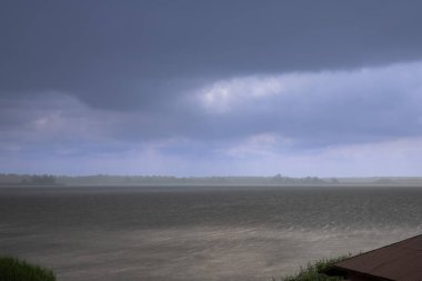 Büyük bir göl üzerinde sağanak yağış, üzerinde kara fırtına bulutları ve ön planda sazlıklar var. Düşük görüş mesafesi ve sisli ufku olan dramatik bir manzara..