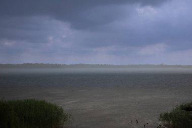 Kara yağmur bulutları ve gölün üzerine sağanak yağış. Görünür sağanak yağış ve rüzgarın, su yüzeyinde doku oluşturduğu fırtınalı bir hava. Fırtınadan önce kırsal doğa manzarası.