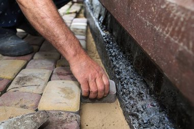 Su geçirmez bir temel duvarı boyunca kaldırım taşları döşeyen bir adamın yakın çekimi. Açık hava inşaatı, asfalt döşeme ve ev geliştirme kavramı.