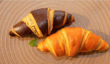 İki taze kruvasan, biri klasik altın ve diğeri siyah çikolata, dekoratif bir paspasın üzerinde yatıyor. Sıcak bir ortamda kahvaltı ya da tatlı için ideal..