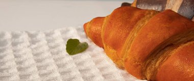 Yeşil nane yaprağıyla beyaz desenli mutfak havlusunda altın kruvasan. Yumuşak sabah ışığı sıcacık ve lezzetli kahvaltı düzenini vurgular.