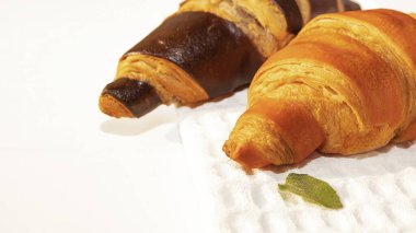 Küçük bir nane yaprağıyla servis edilen yumuşak beyaz peçetede taze altın ve çikolata kruvasanları. Doğal ışık ile minimalist besin bileşimi.