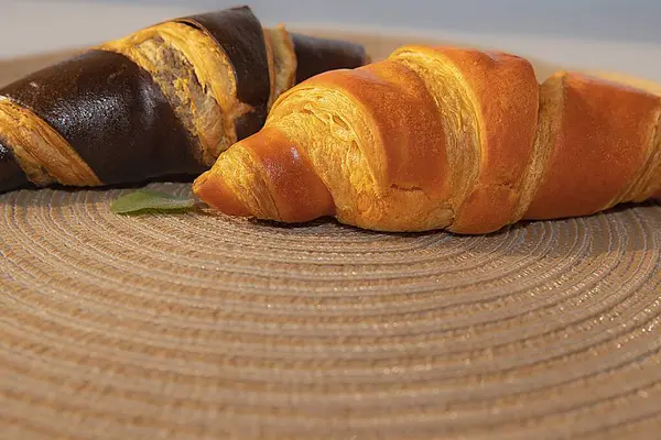 İki taze kruvasan, biri klasik altın ve diğeri siyah çikolata, dekoratif bir paspasın üzerinde yatıyor. Sıcak bir ortamda kahvaltı ya da tatlı için ideal..