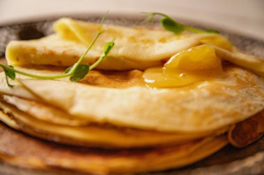 Altın kayısı reçelli ince krep ve antika bir tabakta mikro yeşillikler. Doğal ışık konforlu, ev yapımı kahvaltı atmosferini arttırır..