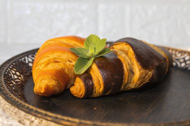 İki yeni pişmiş kruvasan, biri altın ve biri çikolata kaplı, naneli siyah bir tabakta. Kahvaltı ya da pastane için şık yemek sunumu.