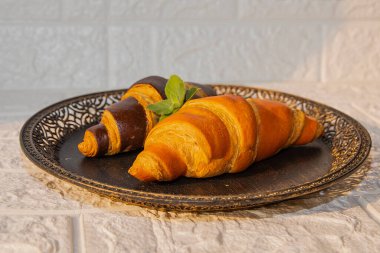 İki yeni pişmiş kruvasan, biri altın ve biri çikolata kaplı, naneli siyah bir tabakta. Kahvaltı ya da pastane için şık yemek sunumu.