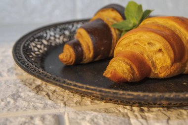 İki yeni pişmiş kruvasan, biri altın ve biri çikolata kaplı, naneli siyah bir tabakta. Kahvaltı ya da pastane için şık yemek sunumu.