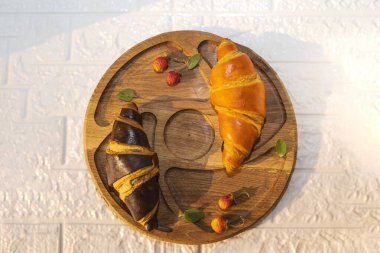 İki kruvasan, bir çikolata ve bir altın, taze yaban çileği ve nane yapraklı ahşap bir tahtanın üzerinde. Düz konumda sıcak güneş ışığı ve doğal dokularla besin düzenlemesi.