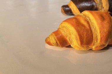 Parlak bej yüzeyde altın ve çikolatalı kruvasanların yakın çekimi. Doğal ışık altında lezzetli taze hamur işleri, fırın ya da kahvaltı temaları için ideal..