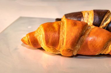 Parlak bej yüzeyde altın ve çikolatalı kruvasanların yakın çekimi. Doğal ışık altında lezzetli taze hamur işleri, fırın ya da kahvaltı temaları için ideal..