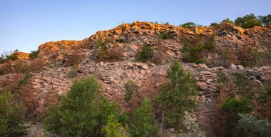 Engebeli taş dokularıyla görkemli kayalık yamaçlar ve sıcak akşam güneşiyle aydınlatılmış dağınık yeşil ağaçlar. Açık gökyüzü ve doğal manzara seyahat, jeoloji ve açık hava temaları için ideal.