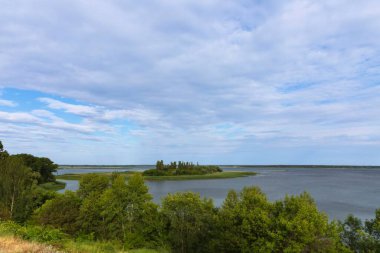 Yeşil adası ve bulutlu gökyüzü olan geniş bir nehir manzarası. Huzurlu doğa manzarası, yaz seyahati ve rahatlama konsepti.