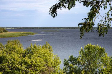 Geniş nehir, yeşil ağaçlar ve bulutlu bir gökyüzünün altında yapraklarla çerçevelenmiş bir tekne. Huzurlu yaz manzarası ve seyahat konsepti.