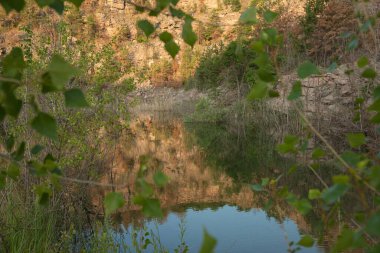 Sakin bir göl kayalık kayalıkları ve parlak bir gökyüzünün altında berrak mavi sudaki bahar ağaçlarını yansıtıyor. Doğa, seyahat ve rahatlama temaları için ideal.