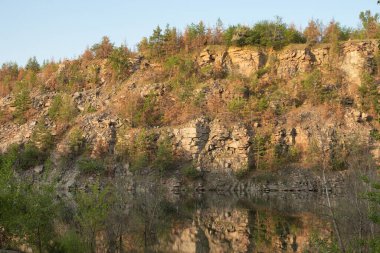 Sakin bir göl kayalık kayalıkları ve parlak bir gökyüzünün altında berrak mavi sudaki bahar ağaçlarını yansıtıyor. Doğa, seyahat ve rahatlama temaları için ideal.