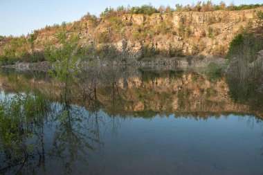 Kayalık kayalık, yeşil ağaçlar ve mavi gökyüzü ile taşan sakin ve berrak bir taş ocağının sularını yansıtarak seyahat ve rahatlama temaları için huzurlu bir doğa manzarası oluşturdu..