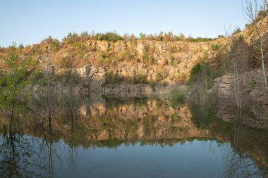 Ağaçlı kayalık, gün batımında taşan taşocağının sakin sularına yansıyarak seyahat, doğa ve rahatlama temaları için huzurlu bir doğal manzara yaratıyor..