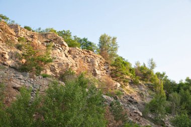 Açık mavi gökyüzünün altında yeşil ağaçları ve çalıları olan kayalık bir uçurum, sıcak güneş ışığına yakalanmış. Doğa, jeoloji, seyahat ve açık hava temaları için ideal.