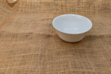 Kırsal çuval bezi kumaşa yerleştirilmiş minimalist beyaz seramik kare kase. Mutfak modelleri, yemek fotoğrafçılığı, tarif blogları ve çevre dostu mutfak tasarımları için ideal..