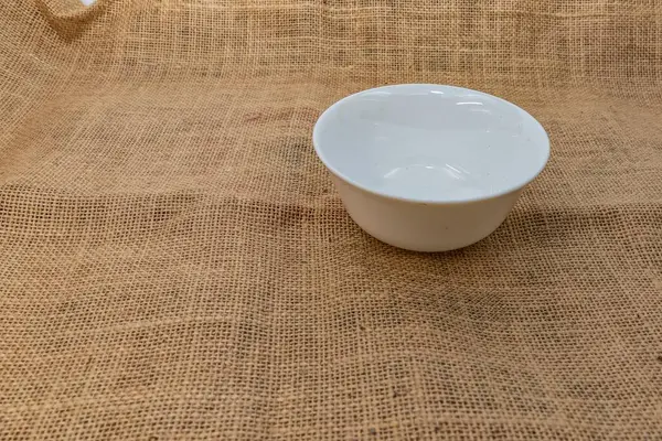 Kırsal çuval bezi kumaşa yerleştirilmiş minimalist beyaz seramik kare kase. Mutfak modelleri, yemek fotoğrafçılığı, tarif blogları ve çevre dostu mutfak tasarımları için ideal..