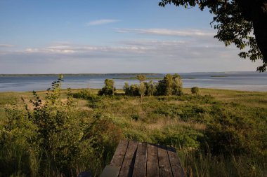 Geniş bir nehir, yeşil çayır ve bulutlu mavi bir gökyüzünün altında dağınık ağaçlarla huzurlu bir yaz manzarası. Doğa, sükunet ve açık hava rahatlığı kavramı.