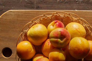 Tahta bir sepetin üzerinde, güneş ışığıyla aydınlatılmış taze, olgun kayısılar. Görüntü, sağlıklı yemek temaları için ideal yumuşak desenli renkli yaz meyvelerini gösteriyor..
