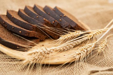 Beyaz bir kâse içinde taze koyu çavdar ekmeği dilimleri ve rustik çuval bezi arka planında buğday kulakları. Sağlıklı geleneksel fırın ürünü.