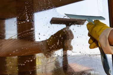 Sabunlu suyla kaplı cam pencereyi silmek için cam sileceği kullanan sarı lastik eldiven giyen birinin yakın çekimi. Temizlik anlayışı, ev işleri ve hijyen..