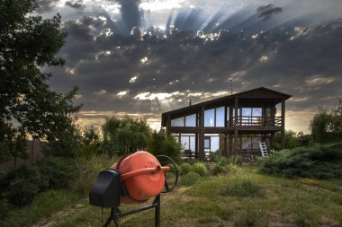 Günbatımında modern bir ahşap evin önündeki inşaat alanında çimento karıştırıcısı. Bulutların arasından geçen güneş ışınlarıyla dramatik bir gökyüzü. Kırsal yaşam, inşaat, mimari ve kırsal çalışma kavramı.