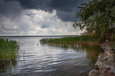 Üzerinde kamış çalıları ve kara fırtına bulutları olan bir göl manzarası. Sağda ahşap bir iskele ve taş setin olduğu görülüyor. Yağmurdan önce huzurlu doğa.