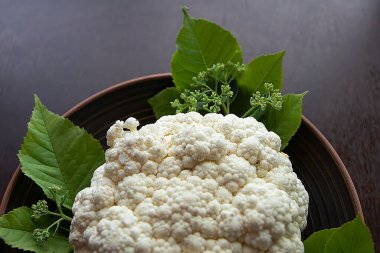 Kıtır beyaz karnabahar yeşil yapraklı, koyu bir tabakta oturuyor, dokusunu ve taze basitliğini vurguluyor..