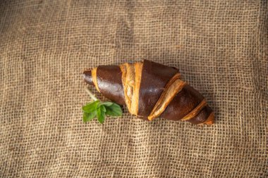 Koyu kahverengi bir kruvasan ve taze fesleğen yaprakları kırsal çuval bezi kumaşına yerleştirilmiş. Dokulu bir kahvaltı sahnesi oluşturuyor.