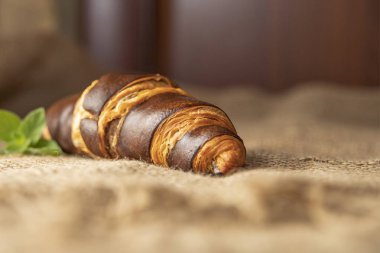 Koyu kahverengi bir kruvasan ve taze fesleğen yaprakları kırsal çuval bezi kumaşına yerleştirilmiş. Dokulu bir kahvaltı sahnesi oluşturuyor.