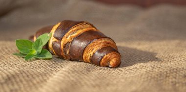 Koyu kahverengi bir kruvasan ve taze fesleğen yaprakları kırsal çuval bezi kumaşına yerleştirilmiş. Dokulu bir kahvaltı sahnesi oluşturuyor.