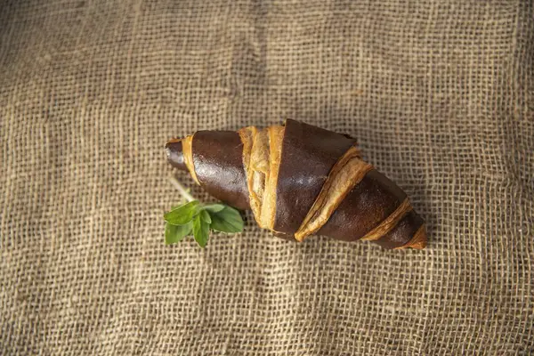 Koyu kahverengi bir kruvasan ve taze fesleğen yaprakları kırsal çuval bezi kumaşına yerleştirilmiş. Dokulu bir kahvaltı sahnesi oluşturuyor.