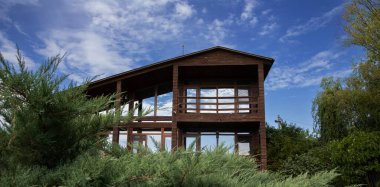 Geniş panoramik pencereleri ve terası olan, temiz mavi gökyüzünün altında ardıç yeşiliyle çevrili, modern ahşap bir ev. Ülke hayatı, mimari ve gayrimenkul kavramı.