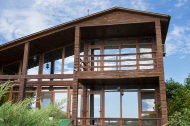 Geniş panoramik pencereleri ve terası olan, temiz mavi gökyüzünün altında ardıç yeşiliyle çevrili, modern ahşap bir ev. Ülke hayatı, mimari ve gayrimenkul kavramı.