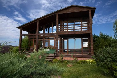 Geniş panoramik pencereleri ve terası olan, temiz mavi gökyüzünün altında ardıç yeşiliyle çevrili, modern ahşap bir ev. Ülke hayatı, mimari ve gayrimenkul kavramı.