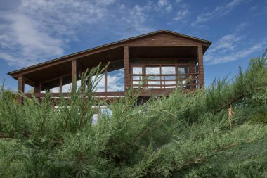 Geniş panoramik pencereleri ve terası olan, temiz mavi gökyüzünün altında ardıç yeşiliyle çevrili, modern ahşap bir ev. Ülke hayatı, mimari ve gayrimenkul kavramı.