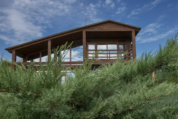 Geniş panoramik pencereleri ve terası olan, temiz mavi gökyüzünün altında ardıç yeşiliyle çevrili, modern ahşap bir ev. Ülke hayatı, mimari ve gayrimenkul kavramı.