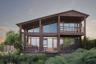 Gün batımında bahçesinde büyük pencereleri olan rahat ahşap bir ev. Tatil yaşam tarzı ve kırsal rahatlama kavramları için mükemmel.