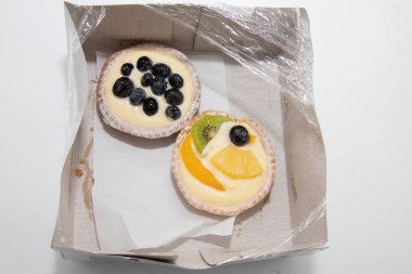 Karton kutuda kremalı iki meyveli tart, biri yabanmersinli, diğeri kivili, şeftalili, ananaslı ve yaban mersinli..