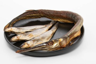 Tütsülenmiş yılan balığı ve beyaz arka plan üzerinde siyah bir tabağa dizilmiş üç küçük balık. Deniz ürünleri ve geleneksel mutfak temaları için mükemmel.