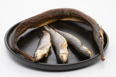 Tütsülenmiş yılan balığı ve beyaz arka plan üzerinde siyah bir tabağa dizilmiş üç küçük balık. Deniz ürünleri ve geleneksel mutfak temaları için mükemmel.