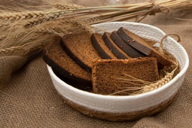 Dilimlenmiş çavdar ekmeğini hasır sepetin içinde kişniş tohumuyla kapat. Buğday kulaklarıyla sade çuval bezi kumaş fırını ve sağlıklı beslenme temasıyla süslenmiş sepetle.