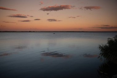 Gün batımında sakin bir göl manzarası. Renkli gökyüzü suya yansıyor. Ufukta küçük bir tekne.