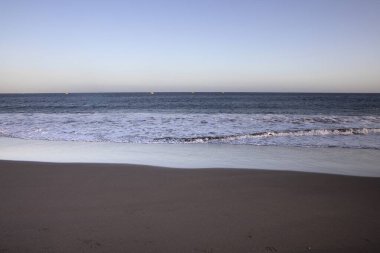 Sakin deniz dalgaları yumuşak ışık yansıması ile kumlu kumsalda yuvarlanıyor. Rahatlamayı, seyahat etmeyi, yazı ve özgürlüğü sembolize eden doğal kıyı arkaplanı.